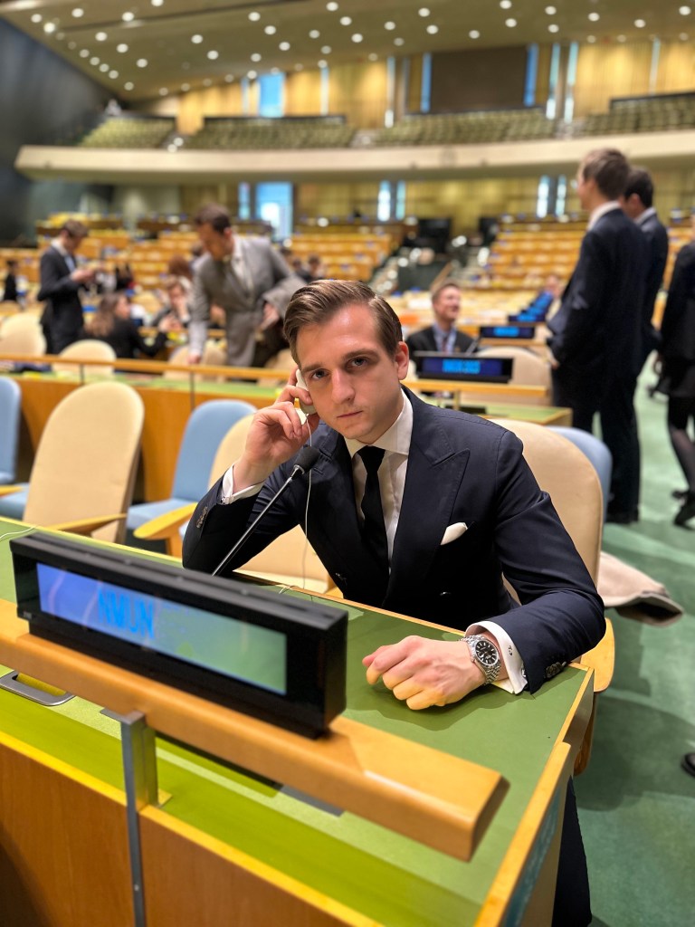 Male delegate in formal suit representing Denmark at the 2023 MUN conference, seated at the United Nations General Assembly hall with a microphone and UN