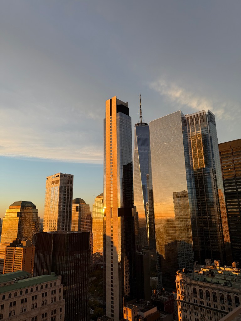 Sunset view of the NYC skyline featuring One World Trade Center and surrounding skyscrapers reflecting golden light.